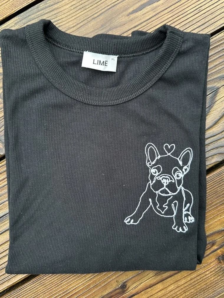 A folded black t-shirt on a wooden table, embroidered with the French bulldog design